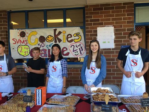 students at bake sale, sign behind reads "SVNMS Cookies for sale"